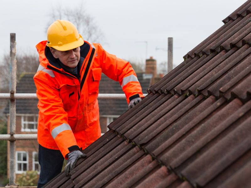 Bilde av en arbeidsmann med hjelm og arbeidsklær på toppen av et stillas. Lener seg på taket og ser på takstenen. 